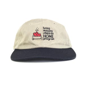 Peut inclure: Casquette de baseball beige avec une visière bleu marine. La casquette présente du texte brodé et un dessin d'un cœur et d'un petit chariot. Le texte se lit "bring missing children HOME program".