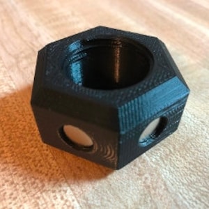 Puede incluir: Un objeto hexagonal negro impreso en 3D con un centro hueco y dos aberturas circulares a cada lado. El objeto tiene una superficie texturizada.
