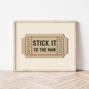 May include: A framed print of a vintage ticket with the text "STICK IT TO THE MAN" printed in black on a brown background. The ticket number 0013472 is printed on both sides of the ticket.