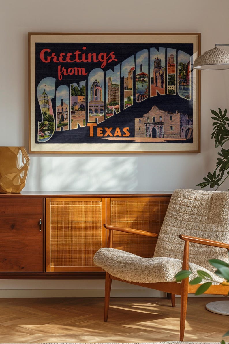 May include: Framed vintage postcard art with the text "Greetings from SAN ANTONIO TEXAS." The artwork features illustrations of San Antonio landmarks. The frame is a light brown color, and the artwork is displayed on a wall above a wooden cabinet and chair.