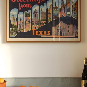 May include: Framed vintage postcard art featuring the words "Greetings from SAN ANTONIO TEXAS" in red and white. The artwork showcases colorful illustrations of San Antonio landmarks. Oranges and a bottle of wine are on the counter.