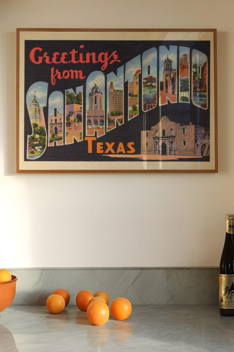 May include: Framed vintage postcard art featuring the words "Greetings from SAN ANTONIO TEXAS" in red and white. The artwork showcases colorful illustrations of San Antonio landmarks. Oranges and a bottle of wine are on the counter.