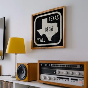 May include: A framed sign with the word "TEXAS", the number "1836", and the word "Y'ALL" in white text on a black background. The sign is in a wooden frame. A yellow lamp and stereo equipment are below the sign.