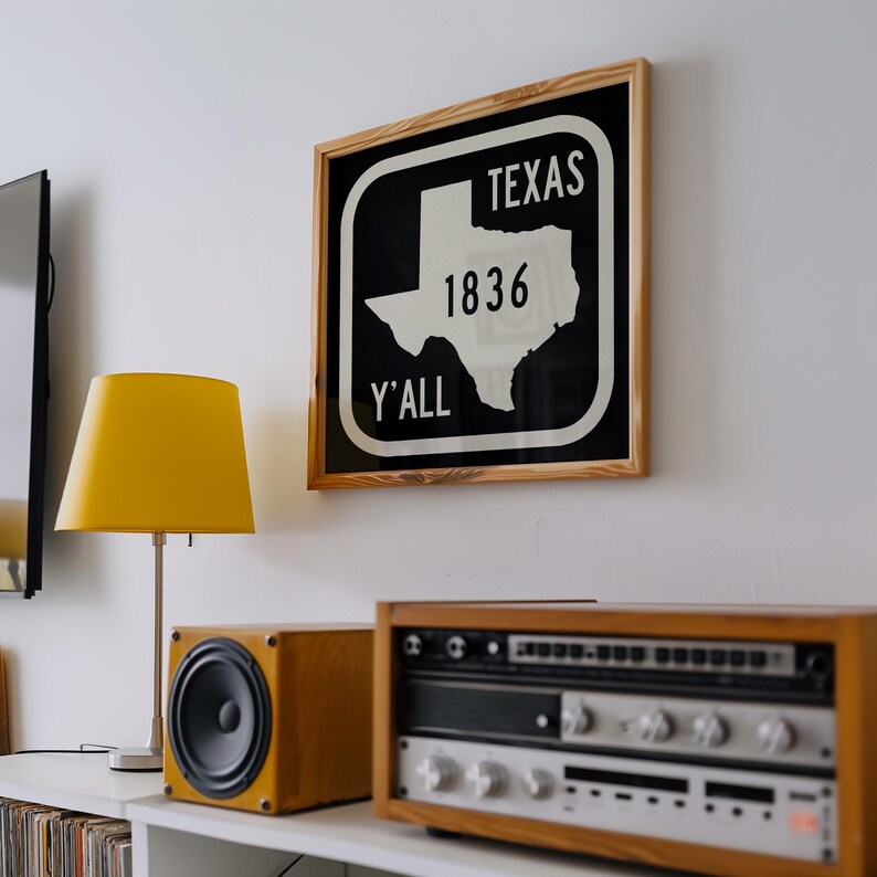 May include: A framed sign with the word "TEXAS", the number "1836", and the word "Y'ALL" in white text on a black background. The sign is in a wooden frame. A yellow lamp and stereo equipment are below the sign.