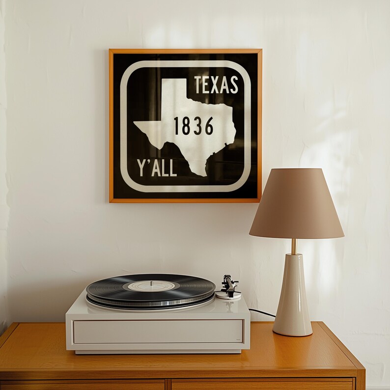 May include: A framed sign with the word "TEXAS" above the outline of the state, the number "1836", and the word "Y'ALL". A white record player with a black vinyl record is on a wooden surface. A beige lamp with a conical shade is also on the surface.