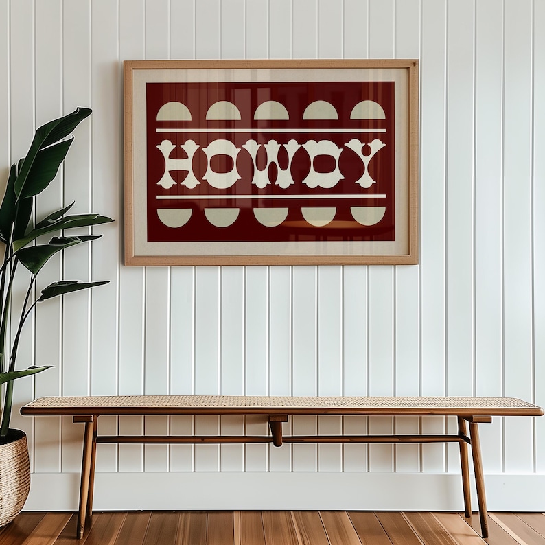 May include: A wooden framed print with a red background and white geometric design. The print says "HOWDY" in white letters. The print is hanging on a white wall above a wooden bench with a woven cane seat.