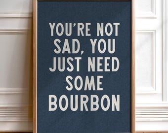 May include: A framed print with the text "YOU'RE NOT SAD, YOU JUST NEED SOME BOURBON" in white letters on a dark blue background. The frame is made of light brown wood. The print is displayed on a wooden floor.