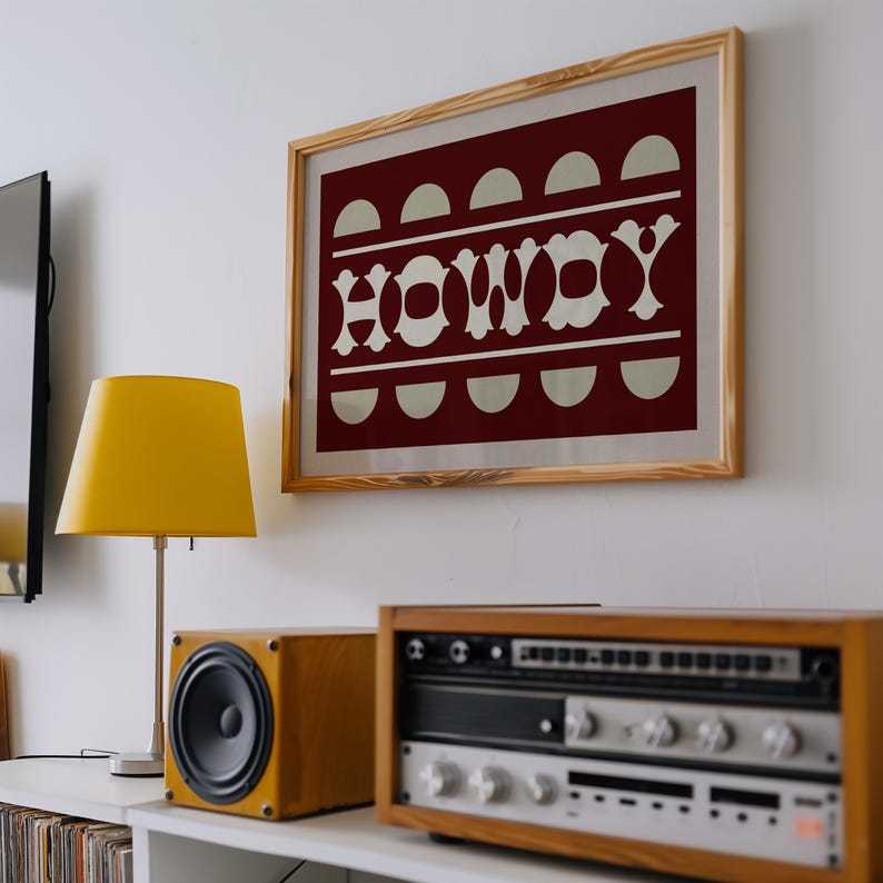 May include: A framed print with a red background and white text that reads "Howdy". The print is hanging on a white wall above a stereo system and a lamp.