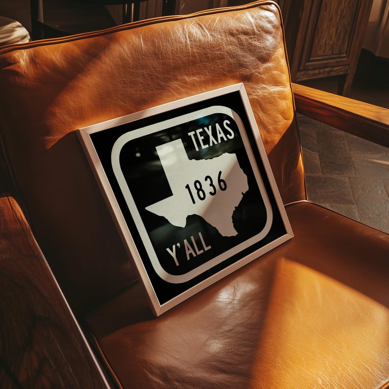May include: A framed sign featuring the word "TEXAS", the number "1836", and the phrase "Y'ALL". The sign displays a white outline of the state of Texas against a black backdrop, within a white frame.