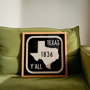 May include: Framed wall art depicting the state of Texas in white against a black backdrop. The text "TEXAS 1836 Y'ALL" is in white. The artwork is enclosed in a light brown wooden frame, suitable for home decor.
