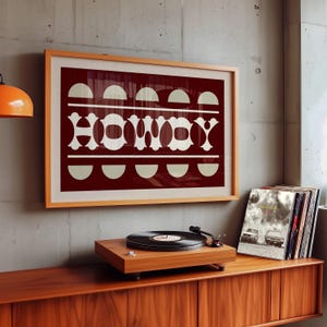May include: A framed print with a red background and white text that says "HOWDY". The text is made up of geometric shapes. The print is hanging on a wall above a record player and a shelf of vinyl records.
