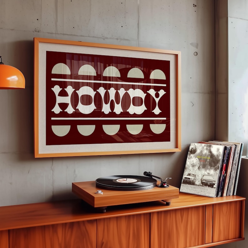 May include: A framed print with a red background and white text that says "HOWDY". The text is made up of geometric shapes. The print is hanging on a wall above a record player and a shelf of vinyl records.