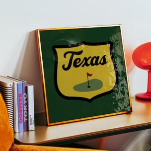 May include: A framed print featuring a shield-shaped design with the word "Texas" in black script. The shield is yellow with a green golf course and a red flag. The background is dark green with a gold frame.