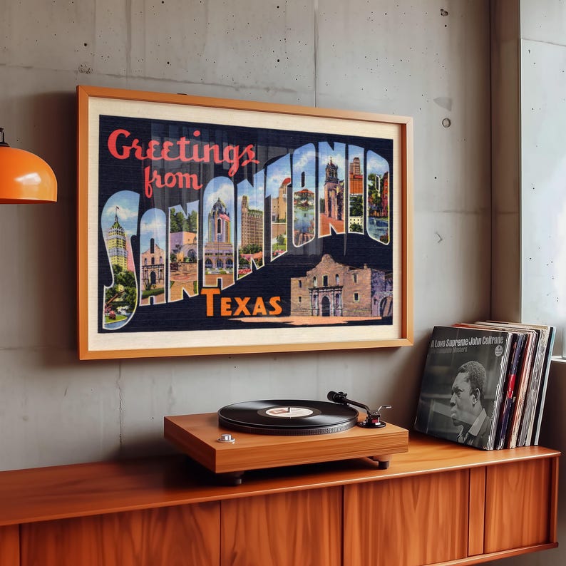 May include: A framed vintage postcard print with the words "Greetings from SAN ANTONIO TEXAS" in a wooden frame. The print is displayed above a wooden record player and a stack of vinyl records.