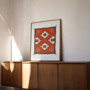 May include: A framed print of a red, white, and black Native American rug with a geometric pattern. The rug is displayed on a wooden cabinet.
