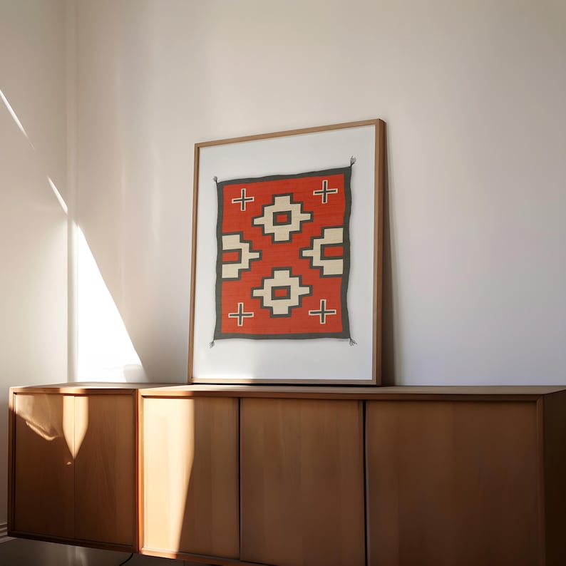 May include: A framed print of a red, white, and black Native American rug with a geometric pattern. The rug is displayed on a wooden cabinet.