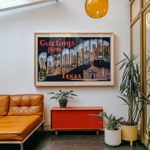 May include: A framed vintage postcard print with the words "Greetings from San Antonio Texas" in red and white. The print features illustrations of San Antonio landmarks. The print is displayed above a red cabinet and a brown leather sofa.