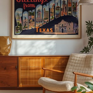 May include: Framed vintage postcard art with the text "Greetings from SAN ANTONIO TEXAS." The artwork features a collage of San Antonio landmarks. The frame is a light brown color. The artwork is hanging above a wooden cabinet and chair.