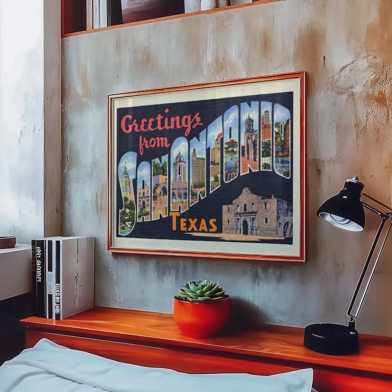 May include: Framed vintage postcard art with the text "Greetings from SAN ANTONIO TEXAS." The artwork features a collage of San Antonio landmarks and buildings. The frame is a warm brown color, and the artwork is displayed on a textured wall.