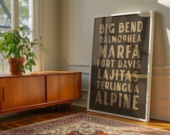 May include: A framed poster with a white border leans against a wall. The poster features the names of Texas locations in gold lettering on a dark background. A wooden cabinet, a potted plant, and a patterned rug are in the foreground.