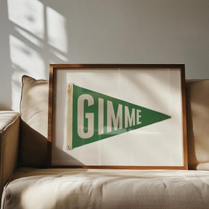 Puede incluir: Un banderín verde enmarcado con la palabra "GIMME" impresa en letras blancas. El banderín está colgado en un sofá beige.
