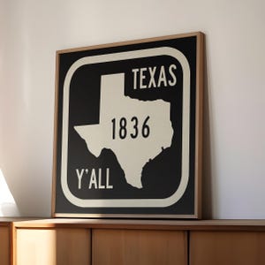 May include: A framed print with a black background and a cream-coloured outline of the state of Texas. The text "TEXAS", "1836", and "Y'ALL" are in white. The frame is made of light brown wood.
