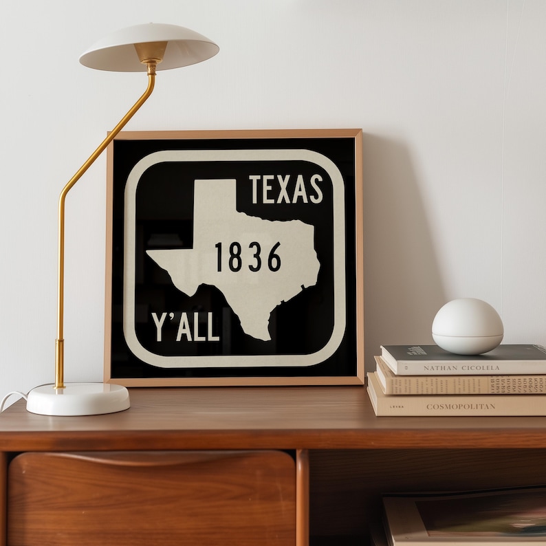 May include: A framed black and white sign with the word "TEXAS" at the top, the number "1836" in the middle, and the word "Y'ALL" at the bottom. The sign features a white outline of the state of Texas. A gold lamp and books are on a wooden surface.