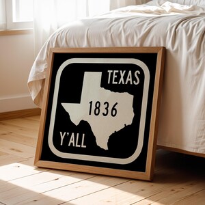 May include: A framed sign featuring the outline of Texas in white against a black background. The text "TEXAS", "1836", and "Y'ALL" are also in white. The frame is made of light-coloured wood. The sign is propped against a bed.