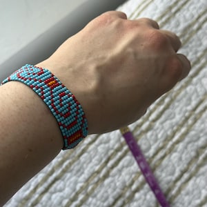 May include: A beaded bracelet with a geometric pattern. The bracelet features small, colorful beads in shades of turquoise, red, and orange. The bracelet is worn on a wrist, showcasing the intricate design and craftsmanship.