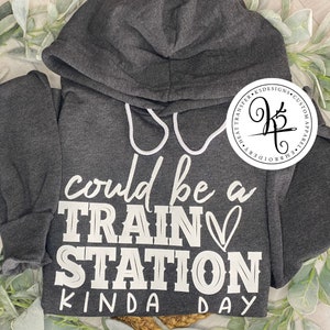 Op de afbeelding: Een donkergrijs hoodie met de tekst "It could be a train station kinda day" in wit gedrukt op de voorkant. De tekst is in een casual, handschriftlijk lettertype.