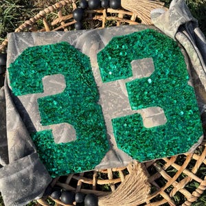 May include: A gray camouflage patterned sweatshirt featuring the number 33 in large, bright green sequin patches. The garment is displayed on a woven basket base decorated with black wooden beads and a jute tassel, showcasing the textured finish.
