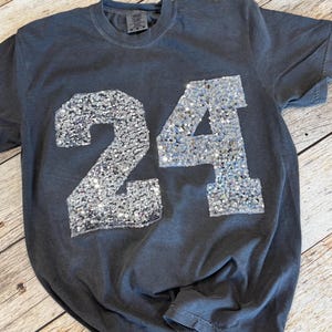 May include: A charcoal gray short-sleeve t-shirt featuring the number 24 on the front. The number is constructed from silver sequins that create a reflective, textured surface. The shirt has a crew neckline and a Comfort Colors brand label.