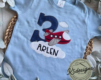 May include: A light blue t-shirt with a navy blue number 3, a red airplane with gray wings, and the name "Arlen" in white letters on a white cloud. The shirt is for a child's third birthday.