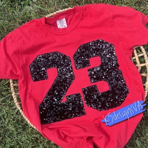May include: A bright red short-sleeved t-shirt featuring the number 23 prominently displayed on the front in large, black, shimmering sequins. The shirt has a crew neckline and a Comfort Colors brand label visible at the inside of the collar.