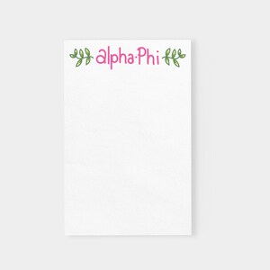 May include: White notepad with a pink "Alpha Phi" text and green leafy vines on the top.