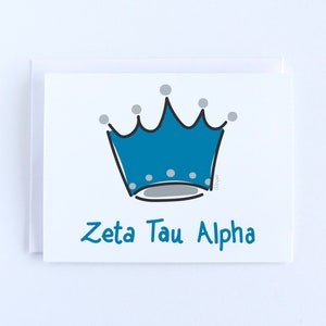 May include: A white greeting card featuring a blue crown illustration with grey circular accents on the points and base. The text Zeta Tau Alpha is printed in blue below the crown. The card is partially tucked into a matching white envelope.