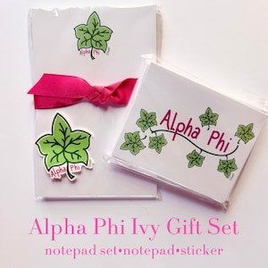 May include: A stationery gift set featuring a white notepad with a green ivy leaf graphic, a matching white notepad with green ivy leaves and the text Alpha Phi, and a die-cut green ivy leaf sticker with the text Alpha Phi, tied with a pink ribbon.