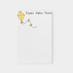 May include: A white notepad with a yellow kite with a black outline and a black and white chevron pattern. The text "Kappa Alpha Theta" is written in black at the top of the notepad.