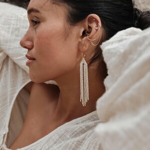 May include: A woman wearing a white shirt with a long, white beaded earring. The earring is made of small, white beads and hangs down to her shoulder.