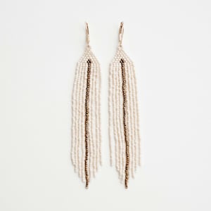 May include: A pair of long, white beaded earrings with a gold accent. The earrings are made of small, white beads and have a single gold line running down the centre of each earring.