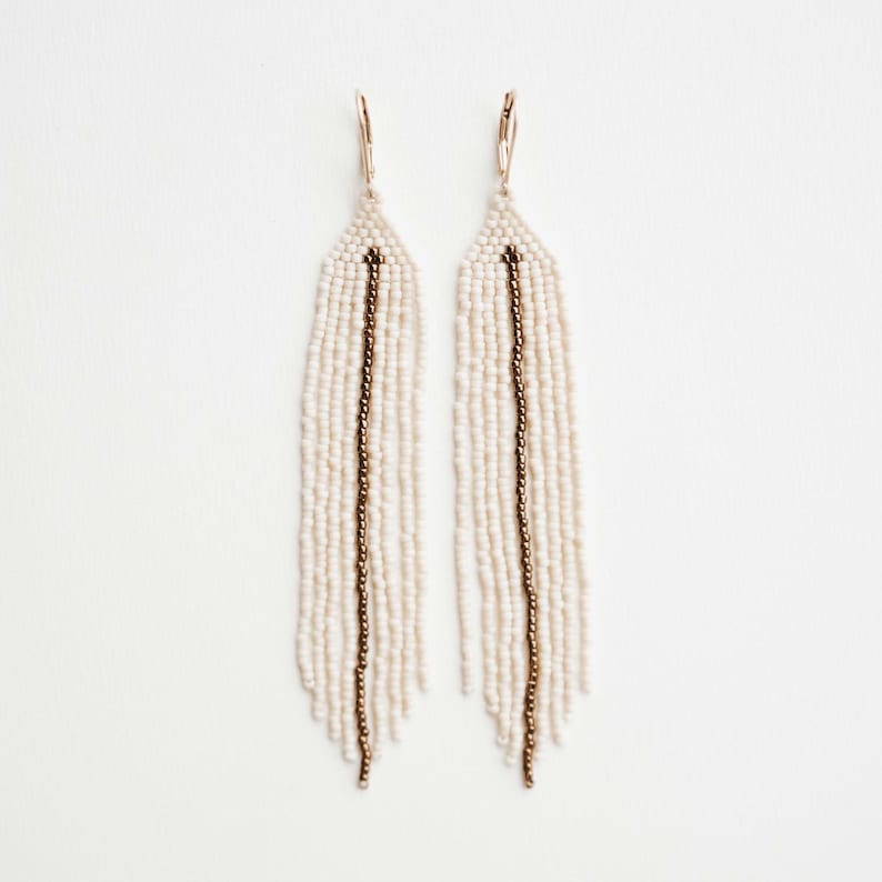 May include: A pair of long, white beaded earrings with a gold accent. The earrings are made of small, white beads and have a single gold line running down the centre of each earring.