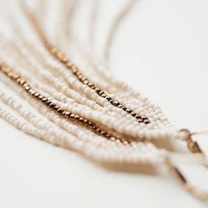 May include: A pair of long, white beaded earrings with gold accents. The earrings are made of multiple strands of beads and have a tassel-like appearance.