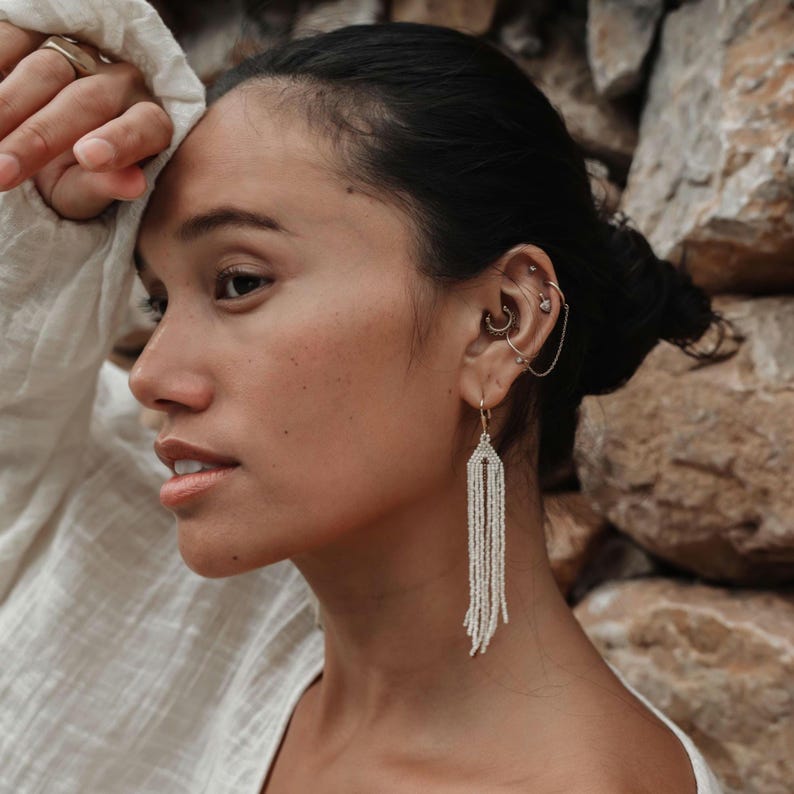 May include: A woman wearing a white shirt with a long, white beaded earring. The earring is made of small, white beads and hangs down to her shoulder.