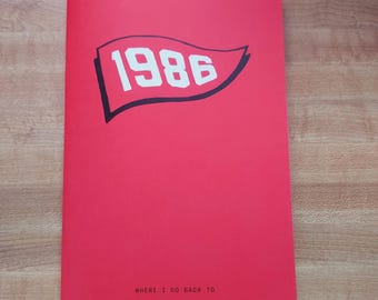 May include: A red booklet with the year "1986" in white, inside a black-outlined pennant shape. The text "WHERE I GO BACK TO" is printed in small, black letters at the bottom of the cover. The booklet is lying on a wooden surface.