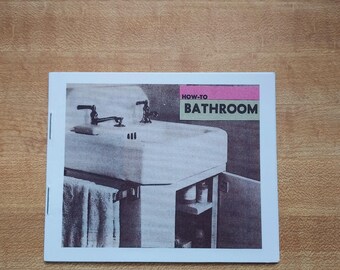 May include: A printed booklet titled "HOW-TO BATHROOM" features a black and white image of a bathroom sink with faucets and a towel rack. The booklet is on a light brown wooden surface.