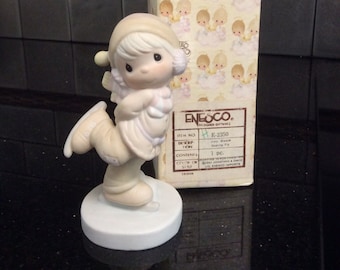 May include: A porcelain figurine of a girl ice skating, wearing a white coat and hat, with a pink scarf. The figurine is on a white base and comes in a box with the text "ENESCO. DESIGNED GIFTS" and "ITEM NO. E-2350".