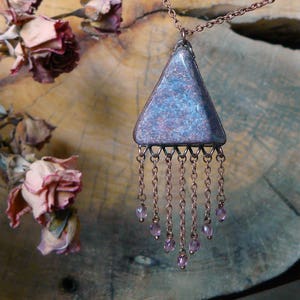 May include: A copper chain necklace with a triangular pendant made of blue and purple speckled ceramic. The pendant has a fringe of copper chains with small purple beads at the ends. The necklace is displayed on a wooden surface with dried roses.