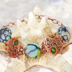 Op de afbeelding: Een koperkleurige armband met ronde en ovale kralen in blauwe, groene en witte tinten. De armband heeft decoratieve koperen schakels en roosvormige filigraan accenten, wat zorgt voor een uniek en artistiek ontwerp.