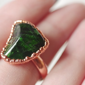 May include: A copper ring with a green gemstone set in a heart shape. The gemstone is surrounded by a copper wire with small beads.