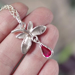 Puede incluir: Un collar de plata con un delicado colgante de mariposa y una piedra preciosa roja colgante. El colgante de mariposa tiene una superficie texturizada y la piedra preciosa tiene forma de lágrima.